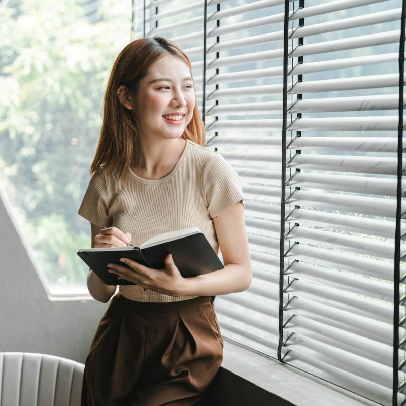 smiling asian woman holding a note in hand and standing near window startup business asian woman onl